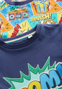 Dwie koszulki T-shirt nałożone jedna na drugą: jedna granatowa z grafiką "BOOM!", a druga w stylu komiksowym z żywymi kolorami i grafikami.