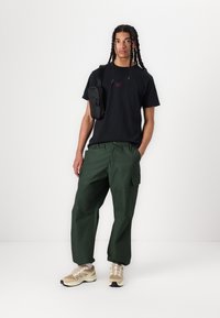 Svart t-shirt med lila logotyp, gröna cargo-byxor med sidofickor och beige sportskor med grå accenter. Justerbar svart axelremsväska.