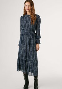 Långärmad, marinblå klänning med en veckad design, texturerad tyg med vertikala metalliska ränder och en åtsittande midja. Svarta knähöga stövlar.