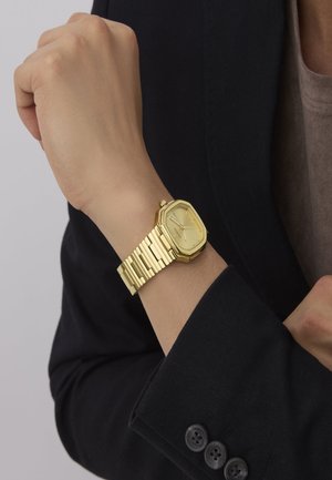Gold octagonal watch with a matching chunky bracelet. The watch face is light gold with silver hour markers, displayed on a wrist.