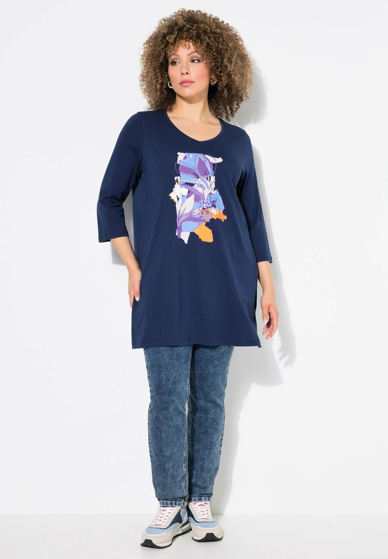 Marineblaue Tunika mit floralem Design in Lila und Orange, mit Dreiviertelärmeln. Tragen Sie sie mit Jeans und Sportschuhen.