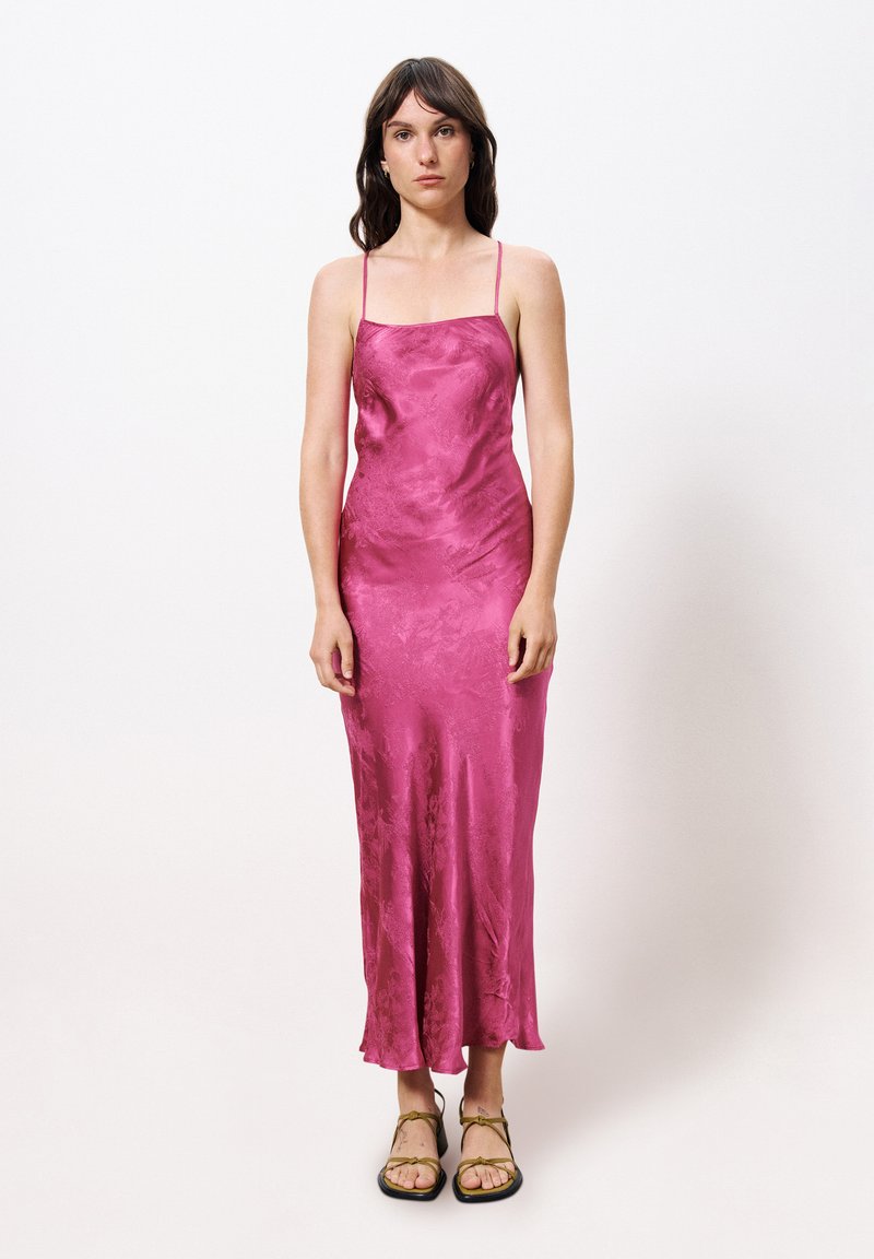 Satin magenta slipdress met dunne bandjes, een passend bovenlijf en een vloeiende zoom. gedragen met bruine sandaaltjes met bandjes. Gladde textuur en minimalistisch ontwerp.