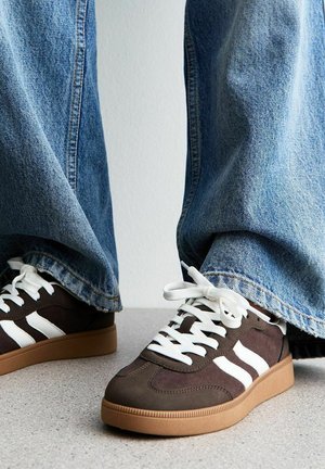 Baskets marron avec des rayures blanches et des lacets, portées avec un jean denim bleu large, debout sur un sol gris moucheté.