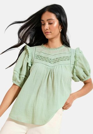 Frau mit langen schwarzen Haaren trägt eine hellgrüne Bluse mit Puffärmeln und Spitzen-Details, kombiniert mit einer weißen Hose vor einem einfarbigen Hintergrund.