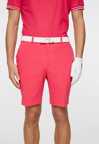 Rosa golfshorts med texturerad tyg, framfickor och ett vitt bälte. Outfitten kompletteras av en vit golfhandske på höger hand.