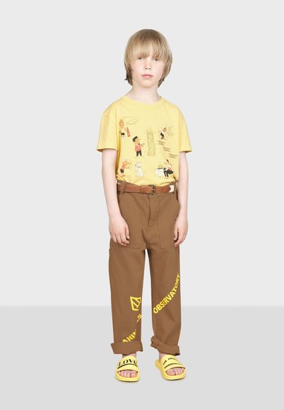 T-shirt jaune avec des graphiques de dessin animé, pantalon marron avec poches latérales et sandales jaunes à enfiler avec texte imprimé. Taille ceinturée visible.