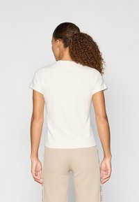 Camiseta de algodón cremosa con mangas cortas, cuello redondo y una forma ajustada. Vista desde la parte de atrás, combinada con pantalones beige que presentan detalles laterales.