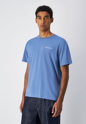 Camiseta de algodón color azul claro con cuello redondo y mangas cortas. Presenta un pequeño logo de "Champion" en rojo y blanco en el pecho y la manga.