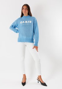 Donna con un maglione blu con testo "PARIS", pantaloni bianchi e tacchi neri, in piedi con una mano sui fianchi su uno sfondo bianco.