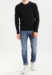 Czarna koszula z długim rękawem i okrągłym dekoltem, zestawiona z jasnoniebieskimi dżinsami i białymi trampkami. Koszula ma małe logo na piersi.