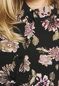 Black floral blouse featuring large pink and beige flowers, green leaves, and black buttons along the front. Soft, textured fabric.