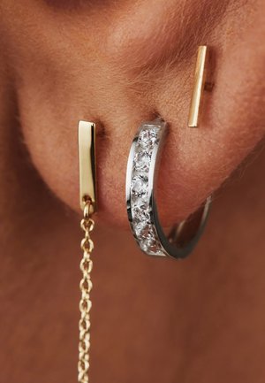 Gold- und Silber-Ohrringe mit klaren Edelstein-Akzenten, die eine Goldstange und eine filigrane Kette verbinden. Glatte Metalloberfläche.