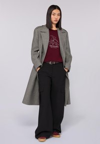 Grauer Wollmantel, burgunderroter Pullover mit Grafikdruck, schwarze weite Cargo-Hosen und bordeauxrote Plattformschuhe mit beigen Sohlen.