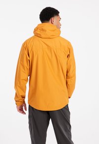 Man in mosterdgele hoodie en donkergrijze broek, met zijn rug naar de camera gekeerd tegen een witte achtergrond.