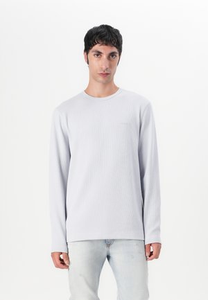 Hellgrau, langärmliges Shirt mit strukturiertem Ribbedesign, runder Ausschnitt und dezentem Logo auf der linken Brust. Kombiniert mit hellen Jeans.