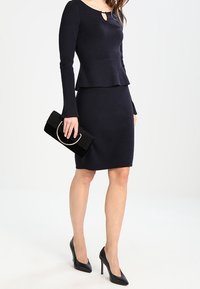 Robe tricotée bleu marine à manches longues avec basque, associée à une pochette noire ornée d'un accent métallique circulaire et à des escarpins noirs à talons aiguilles.