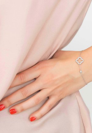 Hand resting on pale pink fabric, wearing a delicate silver bracelet with a quatrefoil charm and red nail polish on fingers.
