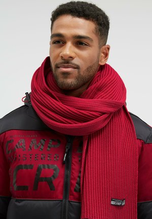 Red ribbed knit scarf wrapped around the neck, made of soft material, featuring a label tag with black text, paired with a black and red jacket.