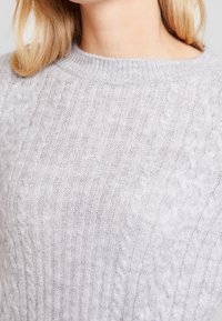 Ljusgrå stickad tröja med ribbad textur och kabelmönster, rund halsringning och mjukt, fluffigt material.