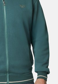Sudadera con cremallera en color teal, de textura suave, con un logo en el pecho y puños y dobladillo acanalados a rayas.