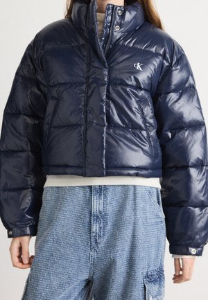 Personne portant une doudoune bleu foncé brillante avec des poches avant et un jean bleu clair texturé, taille haute, avec des poches cargo.