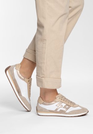 Person, die beige-weiße Turnschuhe trägt, kombiniert mit hochgekrempelten beigen Cordhosen, stehend vor einem schlichten hellen Hintergrund.