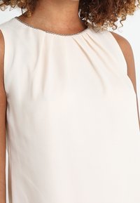 Vestido sin mangas de tela durazno claro con un escote plisado y un acento de cadena plateada a lo largo del cuello. Textura suave.