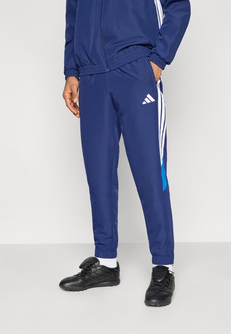 Pantalon de sport bleu marine avec des rayures blanches et un accent bleu clair. Fabriqué en tissu lisse, avec une taille élastique et des chevilles côtelées.