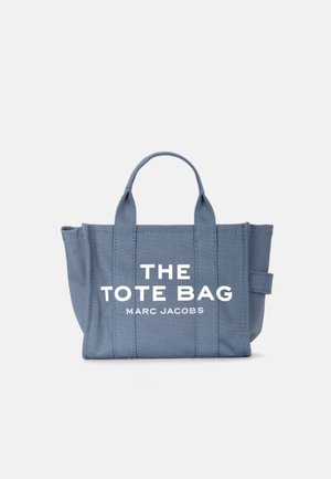 Bolsa de lona azul con dos asas superiores, que presenta un gran texto blanco "THE TOTE BAG" y "MARC JACOBS" en la parte delantera. Textura suave, forma estructurada.
