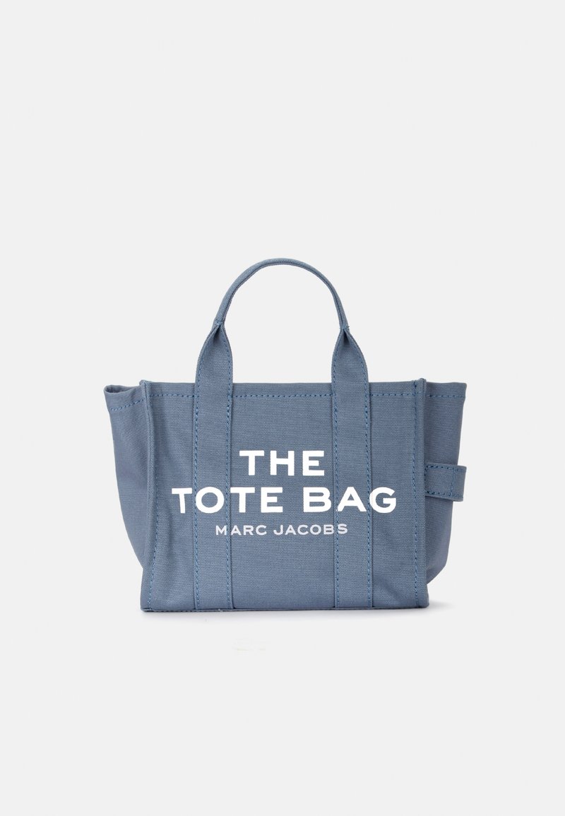 Blå canvas tote-väska med två handtag upptill, med stora vita texter "THE TOTE BAG" och "MARC JACOBS" på framsidan. Slät yta, strukturerad form.