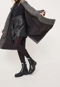 Schwarze Leder Chelsea Boots mit einer robuster Sohle, kombiniert mit einem schwarzen Lederrock und einem grauen strukturierten Mantel, der ein schwarzes Innenfutter hat.