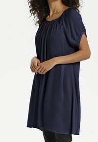 Blouse bleu marine à manches courtes avec une texture plissée, un col rond et un petit détail en forme de goutte à l'avant. Silhouette fluide.