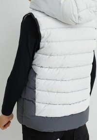 Kamizelka ocieplana z kapturem, biała górna część i szara dolna, pikowana, z kieszeniami oraz gładką zewnętrzną teksturą.