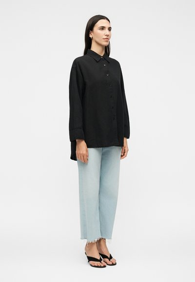 Γυναίκα στέκεται με το πρόσωπο προς τα εμπρός φορώντας ένα φαρδύ μαύρο πουκάμισο με κουμπιά, ανοιχτό μπλε τζιν με ξεφτισμένες άκρες και μαύρα ανοιχτά πέδιλα σε λευκό φόντο.
