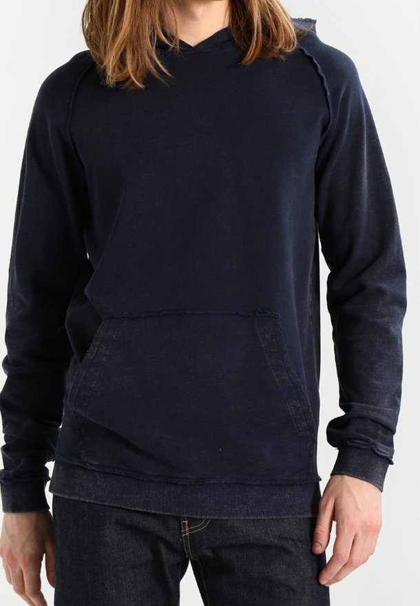 Personne aux longs cheveux bruns portant un sweat à capuche bleu foncé uni et un jean foncé, debout devant un fond clair et uni.