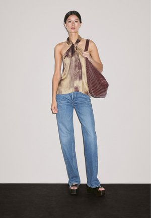 Brun og beige mønstret halterneck top, blå jeans og sorte platform-sandaler; bærer en struktureret burgunder skuldertaske. Hvid baggrund.