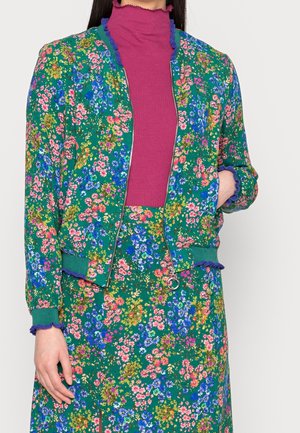 Vrouw die een groen bloemenjasje en bijpassende rok draagt, bedrukt met roze, blauwe en gele bloemen, gecombineerd met een magenta ribgebreide coltrui.