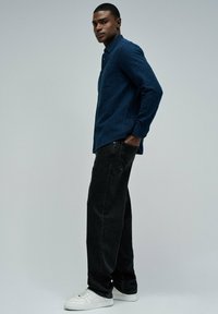 Una camisa oscura de botones azul con un patrón moteado, combinada con pantalones vaqueros negros holgados y zapatillas blancas, creando un atuendo casual.