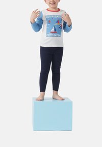Camicia bianca a maniche lunghe con maniche blu, caratterizzata da una stampa nautica. Leggings blu scuro. Il bambino sta in piedi a piedi nudi su un cubo azzurro chiaro.
