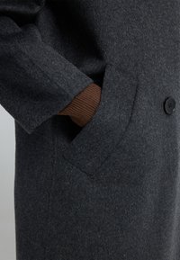 Abrigo de lana gris con cuello de muesca, cierre de un solo botón y bolsillos laterales. La manga revela un detalle acanalado marrón debajo.