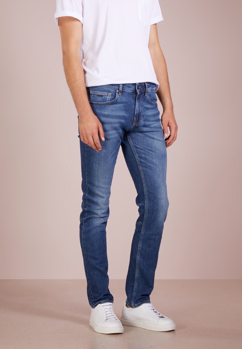 Homme portant un jean slim bleu, des baskets blanches et une chemise blanche à manches courtes, debout devant un fond beige uni.