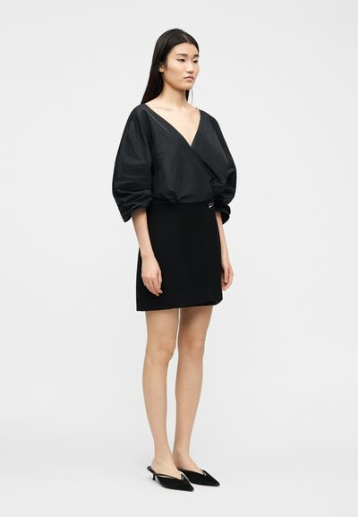 Femme portant un chemisier noir à manches bouffantes et col en V, une mini-jupe noire et des escarpins noirs à bout pointu et bride arrière, debout devant un fond uni.