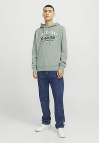 Felpa verde chiaro con stampa grafica frontale, abbinata a jeans di denim blu e sneaker bianche. Presenta un cordoncino e polsini a coste.