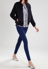 Giacca bomber nera trapuntata, blusa a righe, jeans skinny blu e scarpe con tacco blocco argentate indossate da un modello in movimento.