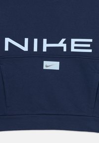 Tmavomodrý sveter Nike s veľkým svetlo modrým logom na prednej strane, štandardný strih, mäkká tkanina, klokanová kapsa a malé logo na vrecku.