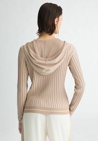 Hoodie beige ajusté avec un côtes verticales, des manches longues et une capuche texturée. Présente une couleur unie et un détail en contraste à l'ourlet.