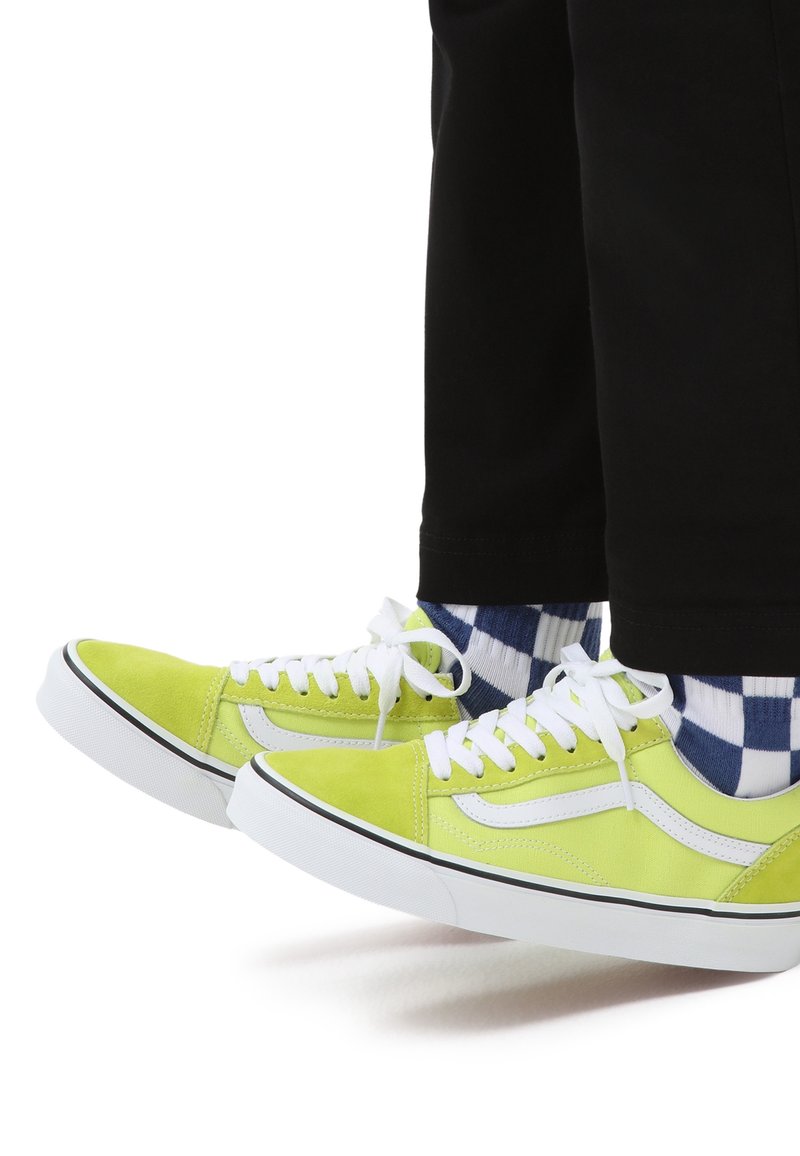 Limegröna sneakers med vita detaljer, i mocka och canvasmaterial, vita skosnören och en svart och blå-rutig strumpa. Svarta byxor syns.