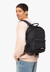 Svart ryggsäck med meshfickor och en framficka med dragkedja, med en logotyp och en rundad design, som bärs över en ljus tröja.