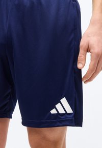 Μπλε σκούρο αθλητικό σορτς σε μηρό ενός ατόμου που δείχνει το λευκό λογότυπο της Adidas κοντά στο τελείωμα και ένα χέρι χαλαρωμένο στο πλάι.