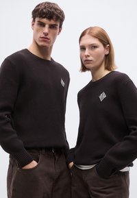 Pulls en maille brun foncé avec une texture côtelée ; chacun présente un petit accent en forme de losange blanc sur la poitrine. Portés avec un pantalon assorti.